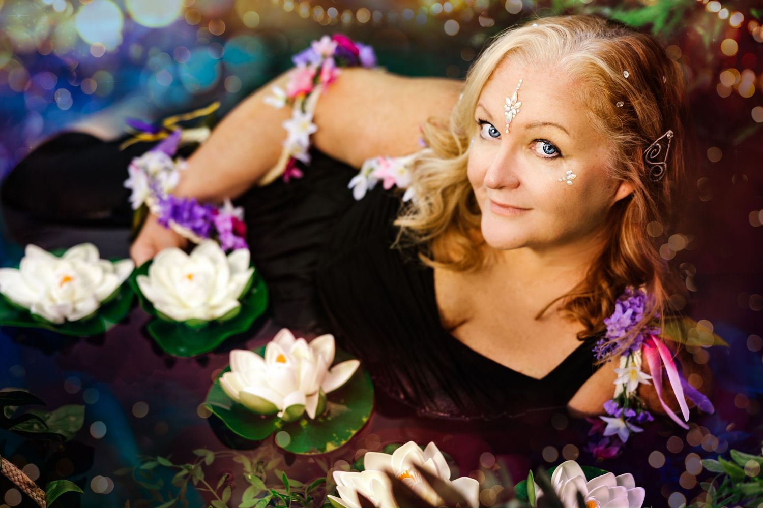A blonde fairy woman with pointed ears lies among white water lilies and purple flowers in an enchanted setting.
