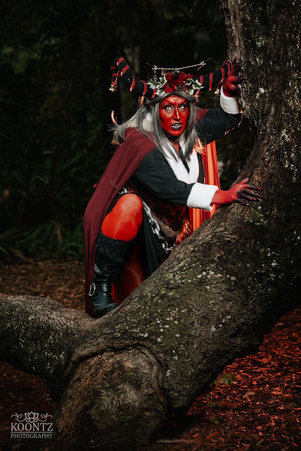 A cosplayer with red body paint, antler crown, and red cape poses dramatically in a large tree in a dark forest.