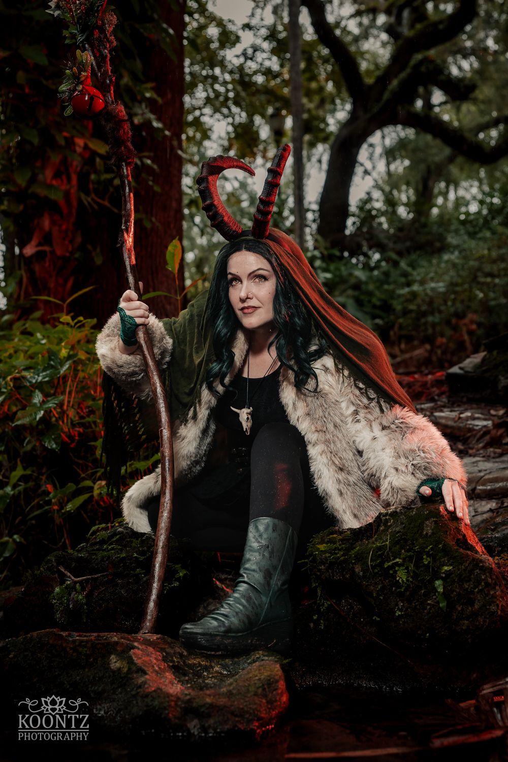Woman in dark fantasy costume with red horns and fur coat holds a staff while seated on mossy rocks.