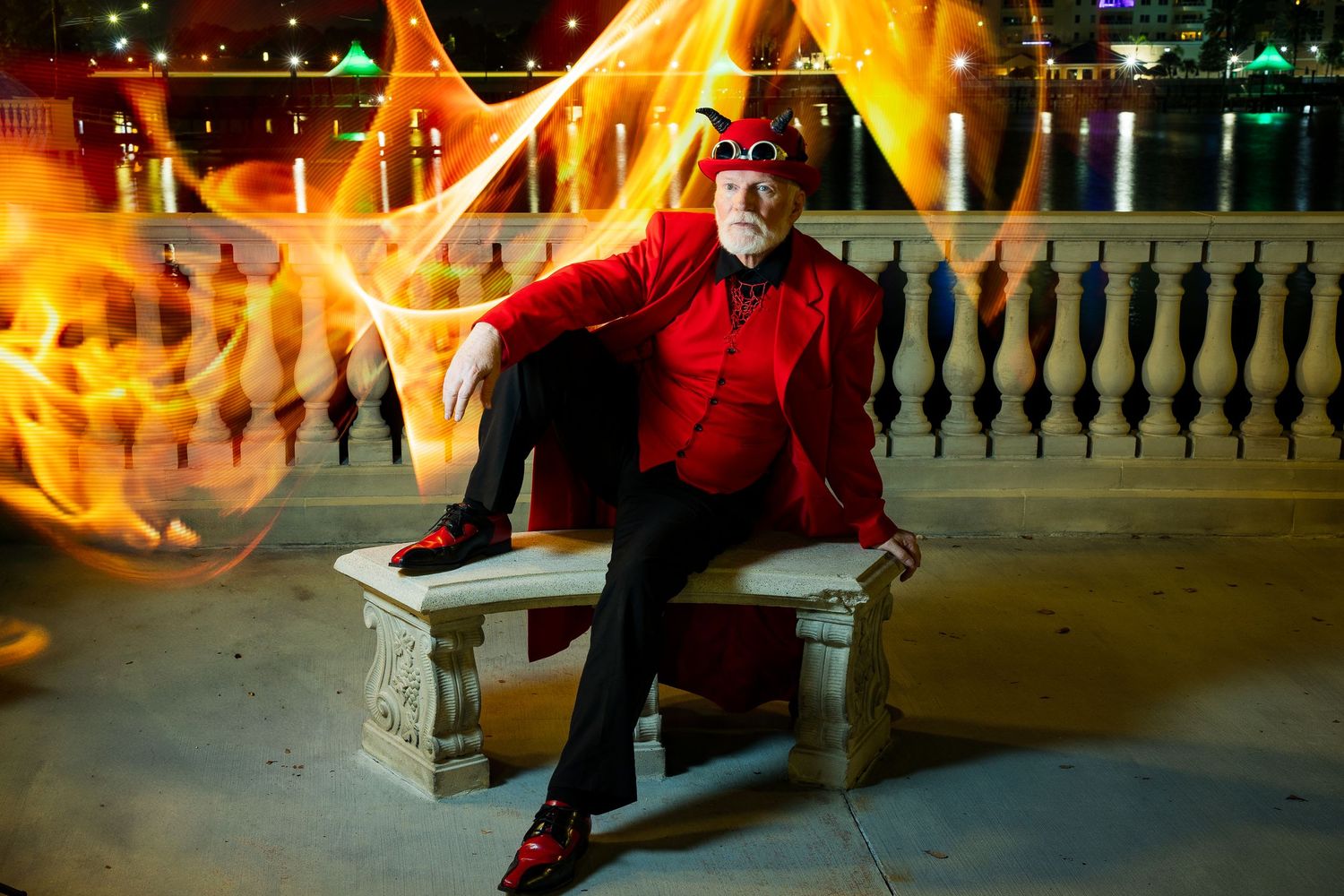 Man in red devil costume sits on stone bench with dramatic orange fire light painting effects at night.