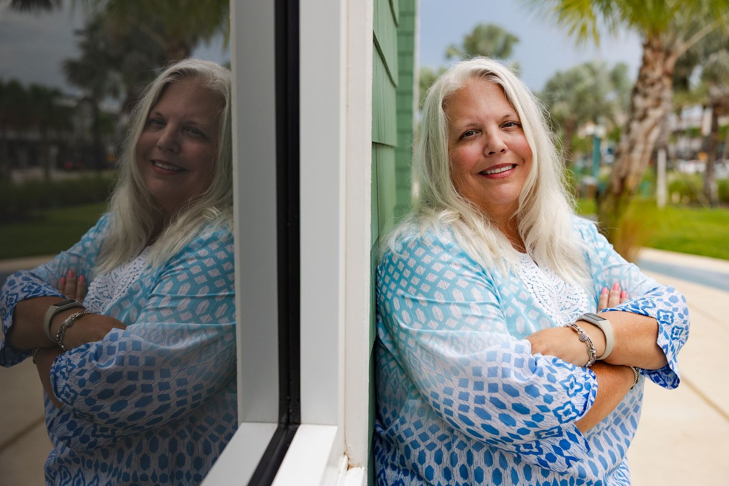 Smiling woman with gray hair in blue patterned top leans against window with reflection visible outdoors.