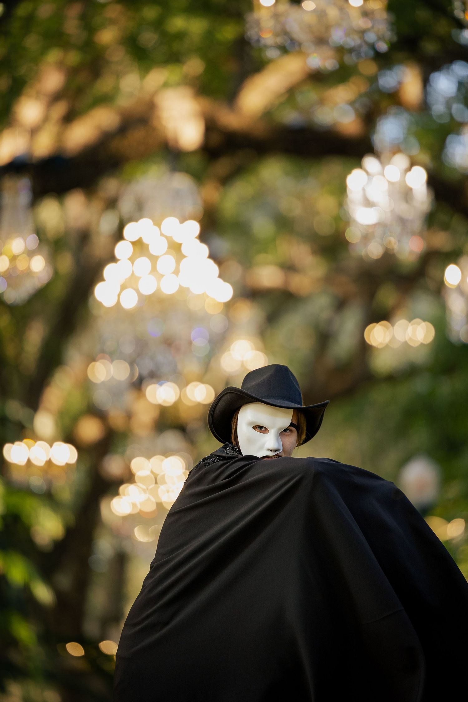 Person in black cape and hat wearing white masquerade mask outdoors with bokeh chandelier lights.