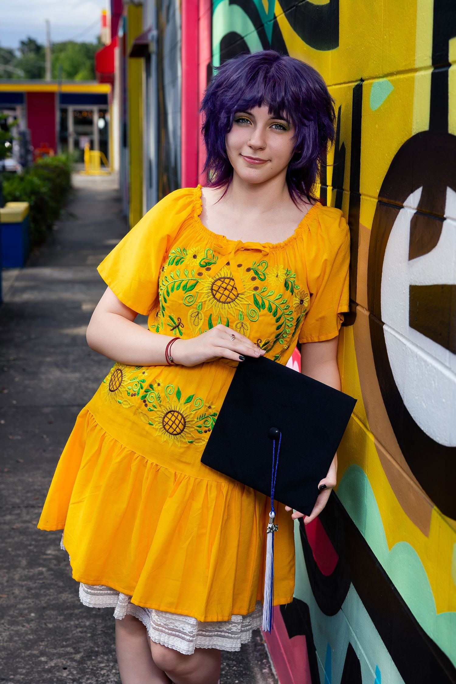 Young woman with purple hair in yellow sunflower dress holding graduation cap, leaning against colorful mural wall.