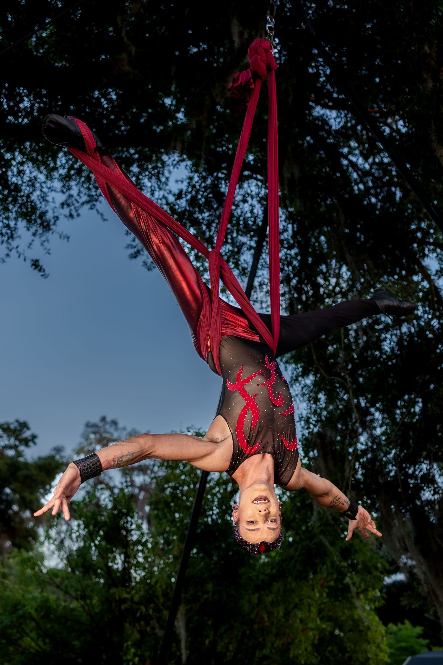 Aerial silk performer in decorative bodysuit hanging upside down from red fabric suspended between outdoor trees at dusk.