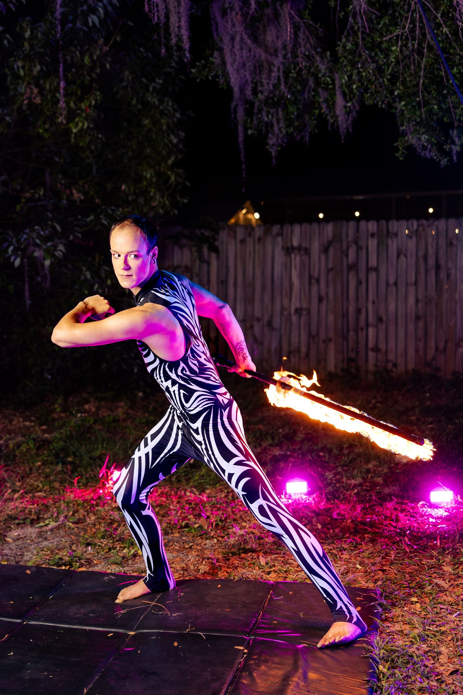 Male fire performer in tribal bodysuit wields a flaming staff in a wide stance at an outdoor night event.