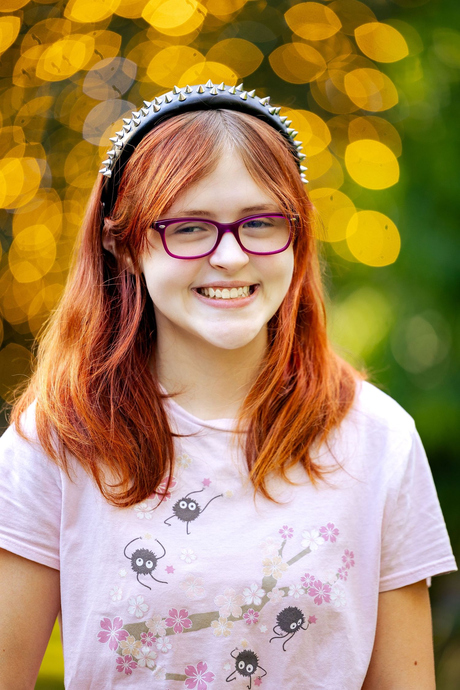 Smiling teenage girl with red hair, purple glasses, and spiked headband wearing a pink graphic tee outdoors.