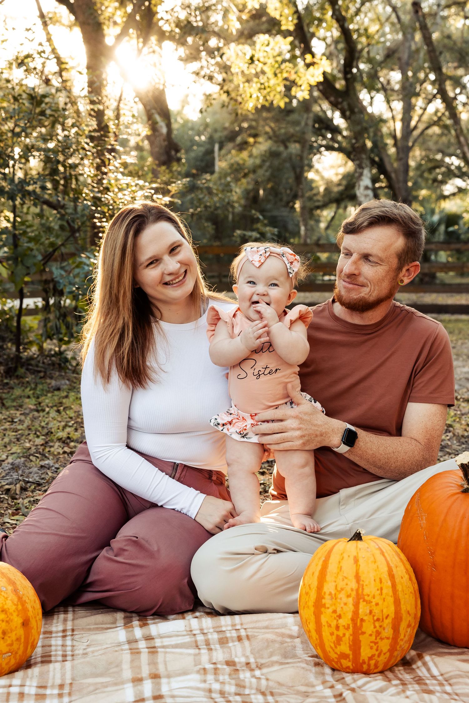 Young family with baby girl sits among pumpkins on a blanket in a sunlit autumn outdoor setting.