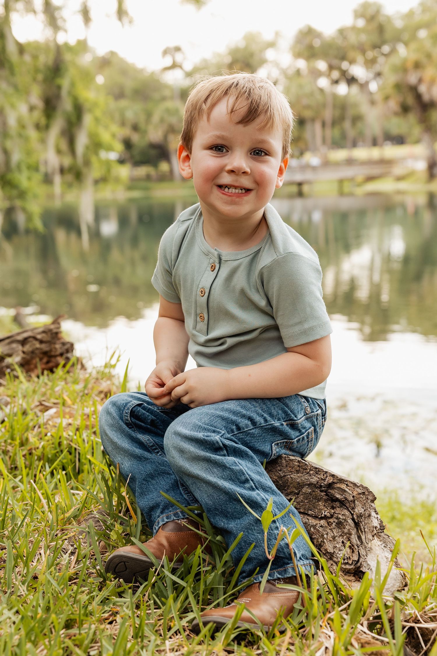 Adorable young boy sitting cross-legged on a log by a peaceful pond, smiling at the camera.