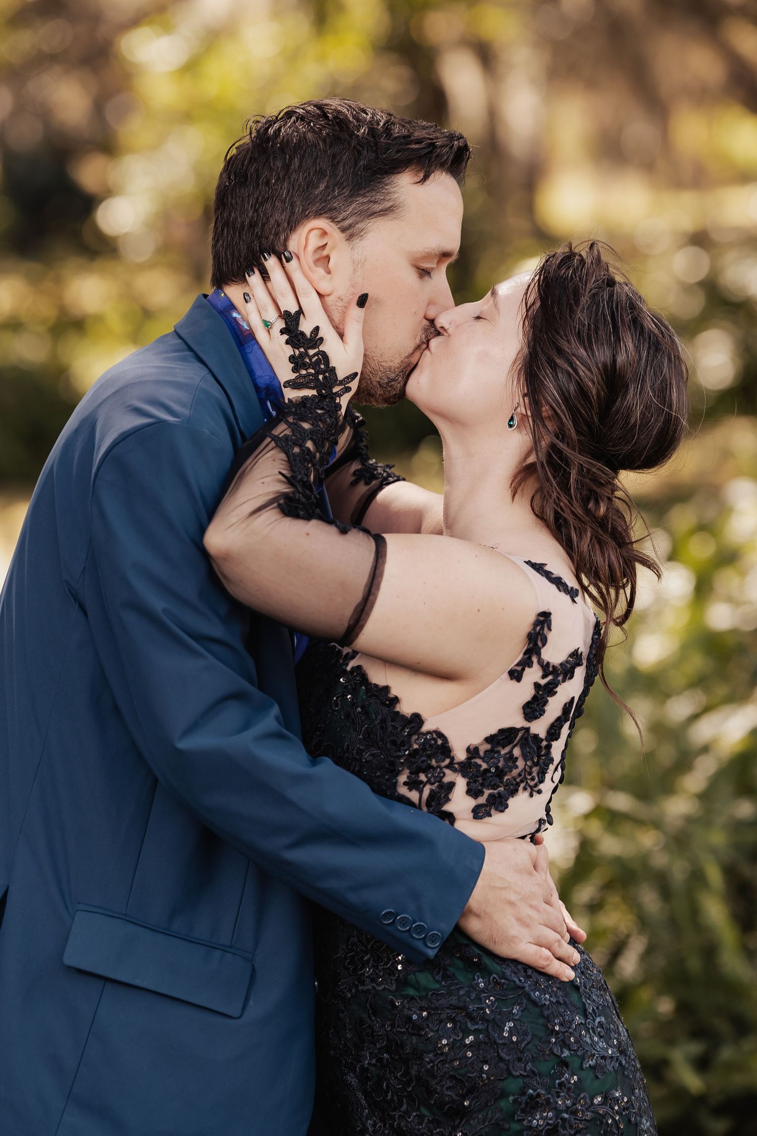 Couple sharing passionate kiss outdoors, groom in blue suit, bride in black lace embroidered gown.