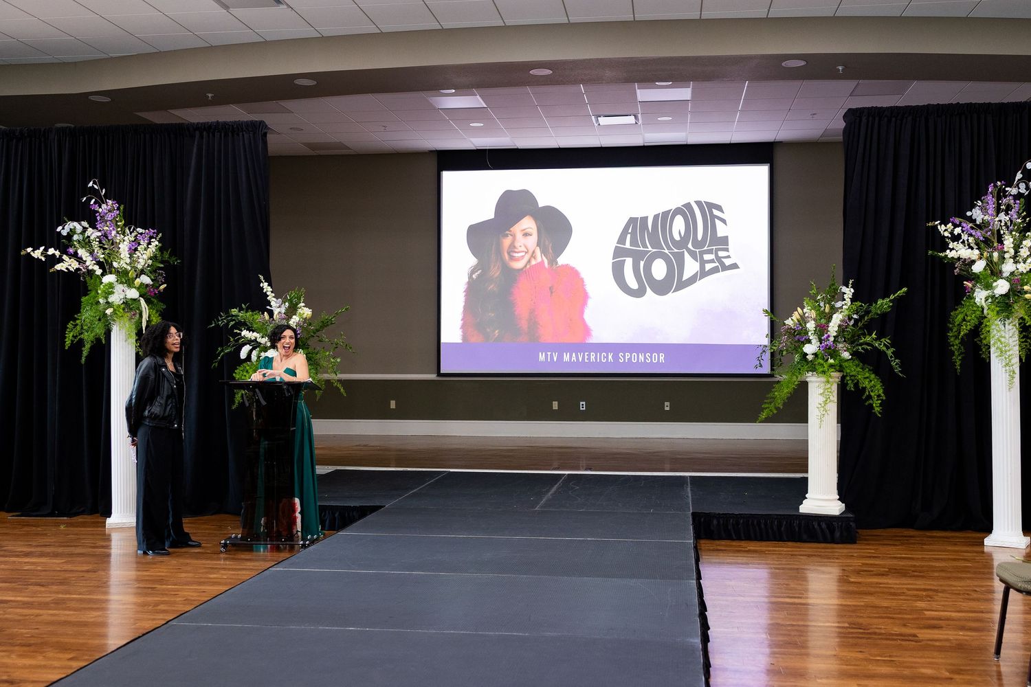 Event stage with large screen displaying Anique Jolee as keynote speaker, flanked by floral arrangements.