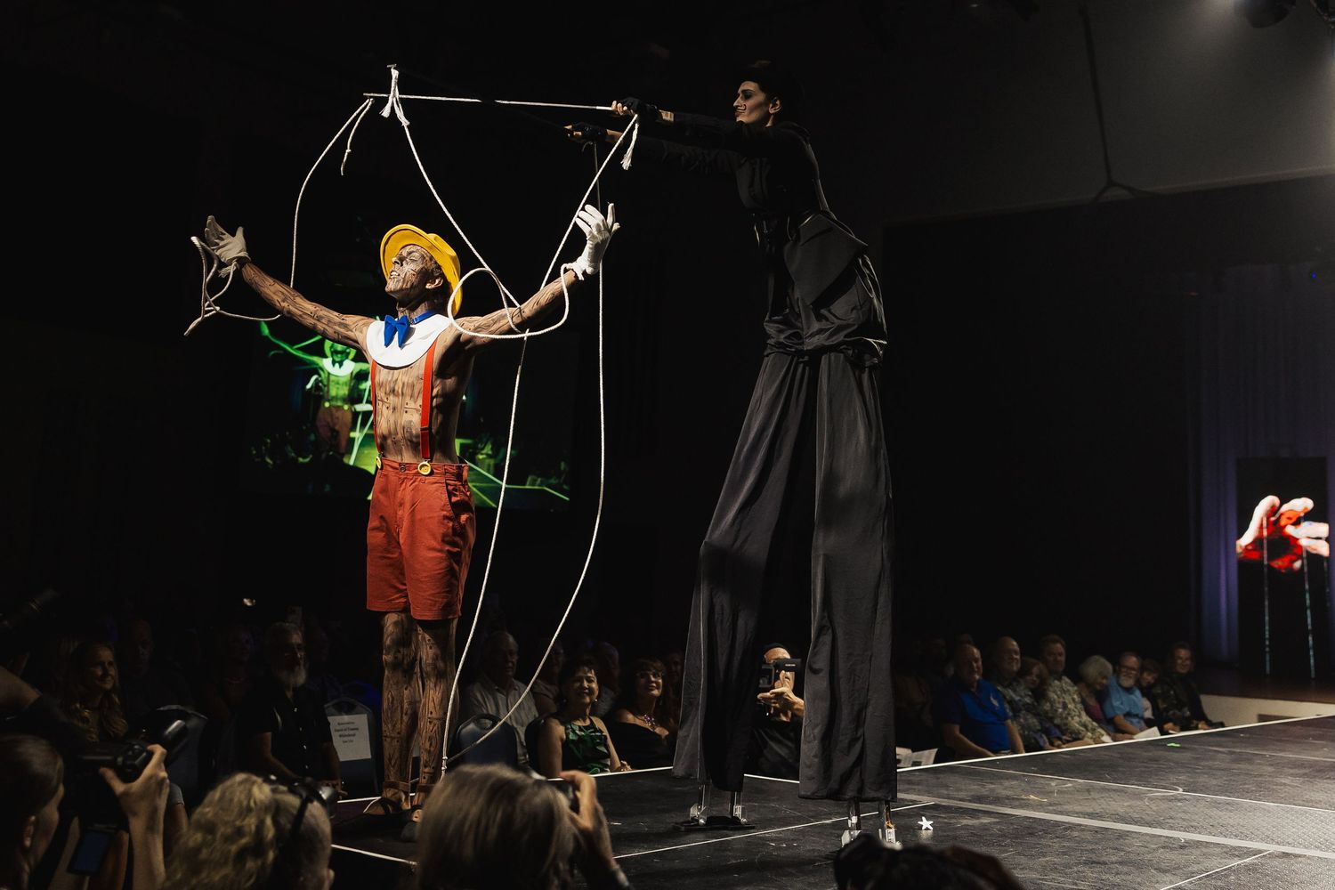 Performer dressed as Pinocchio puppet controlled by tall stilt walker on theatrical fashion runway.