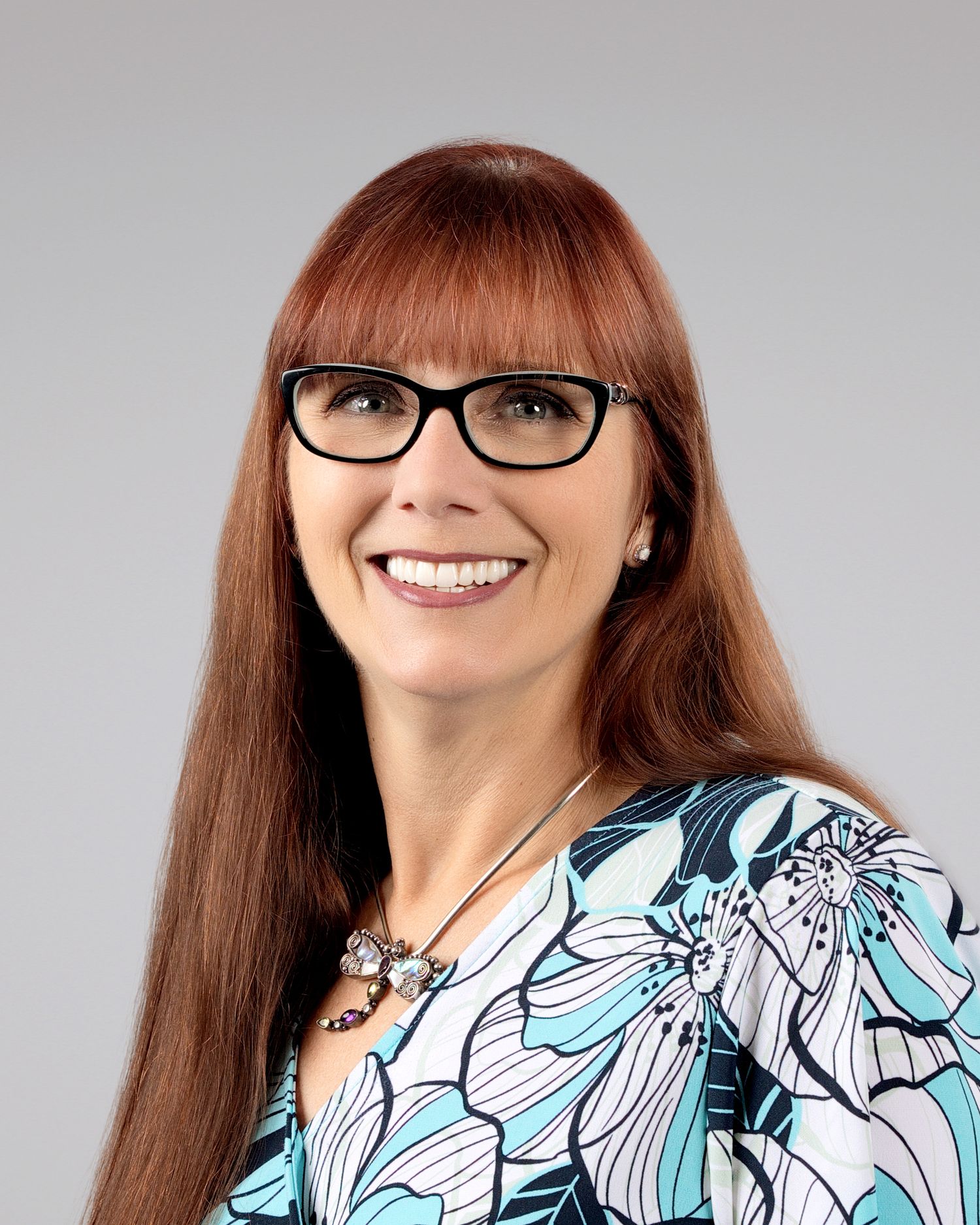 Smiling woman with long red hair and glasses wearing a floral teal and white blouse.