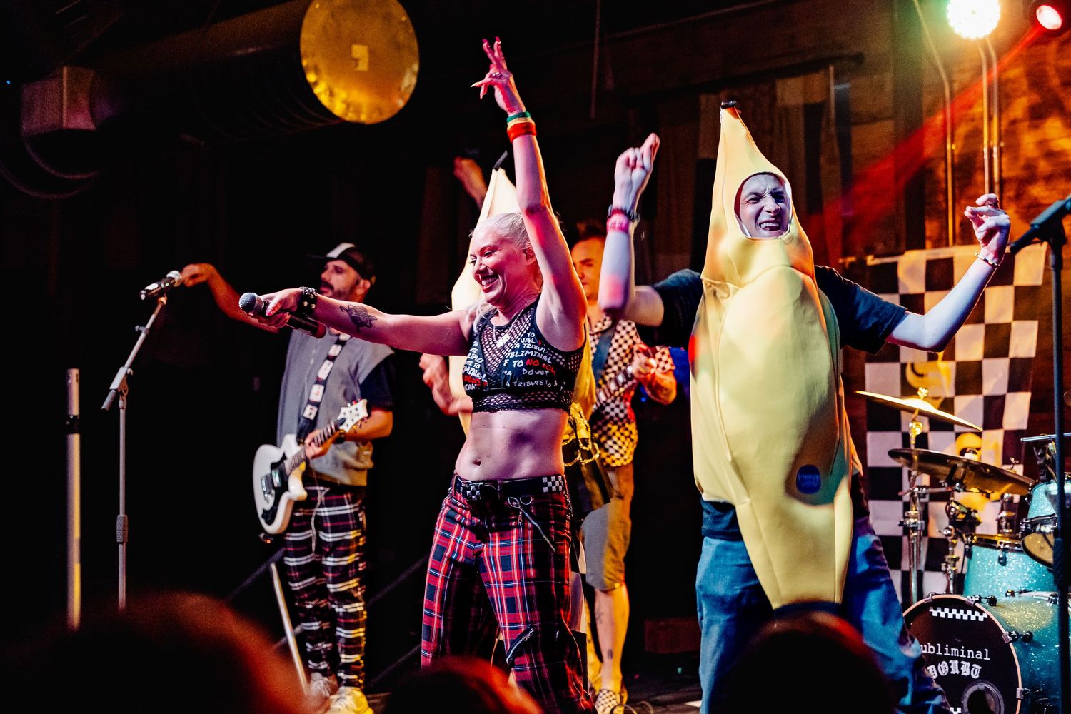 Band performs on stage with members in costumes including a banana suit under checkered backdrop.