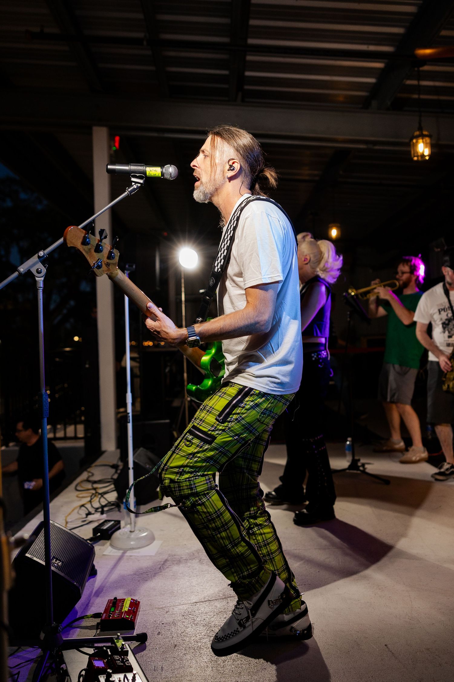 Male bassist in green plaid pants performing energetically on stage at live indoor concert.