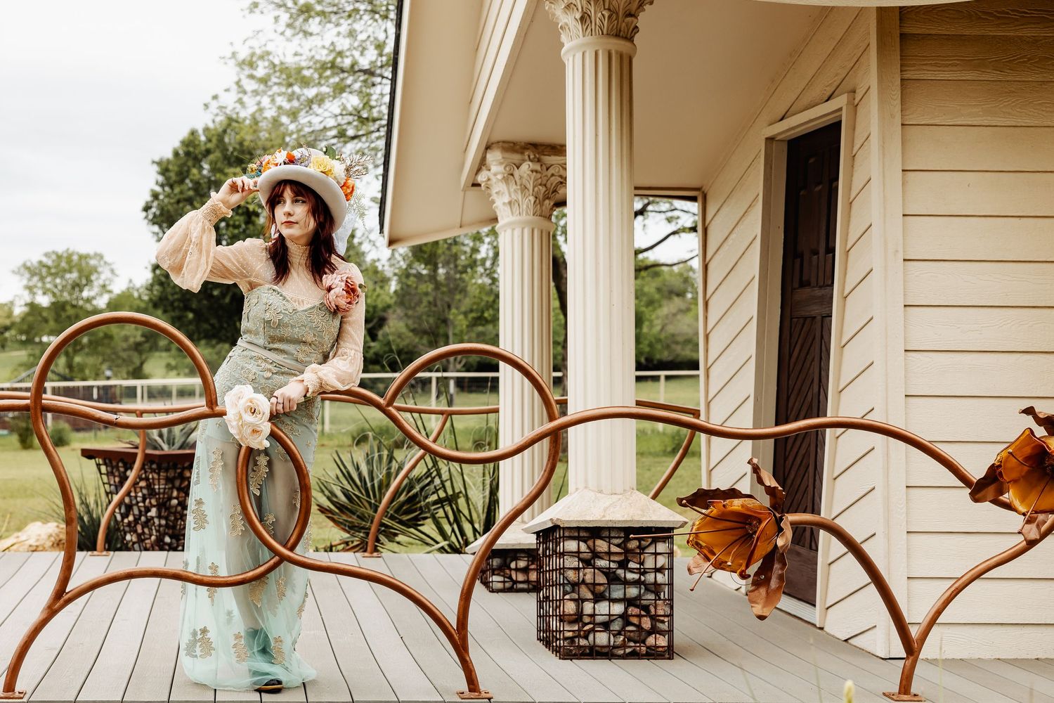 Woman in mint floral dress and straw hat poses on porch with rustic metal garden furniture outdoors.
