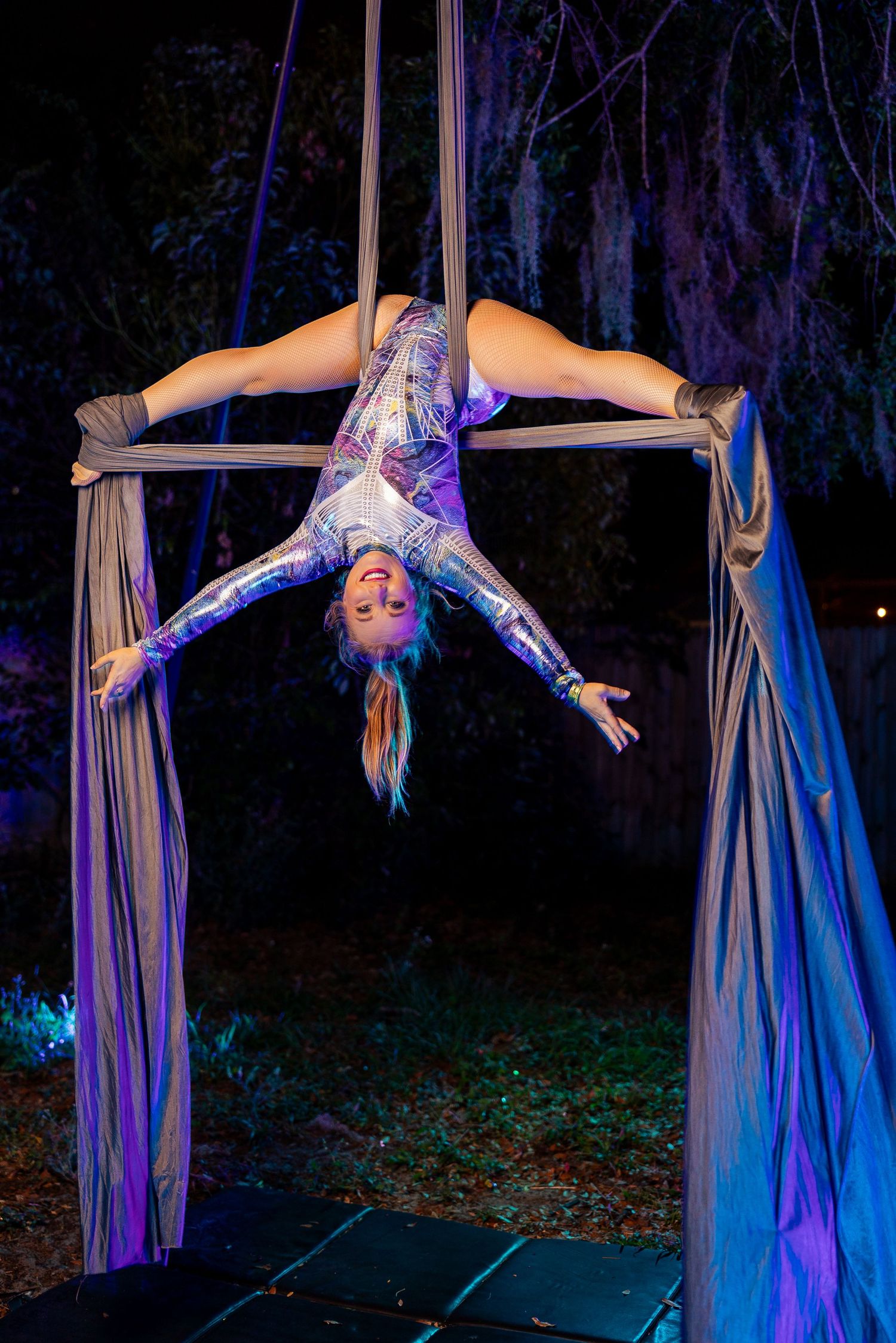 Aerial silk performer in silver costume doing an inverted split on hanging fabric apparatus at night.