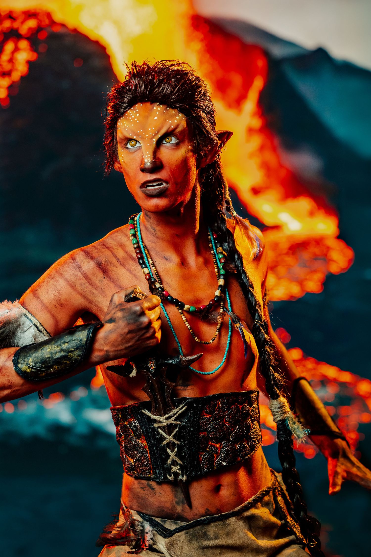 Woman in Avatar-inspired fantasy cosplay with pointed ears and body paint before volcanic eruption backdrop.