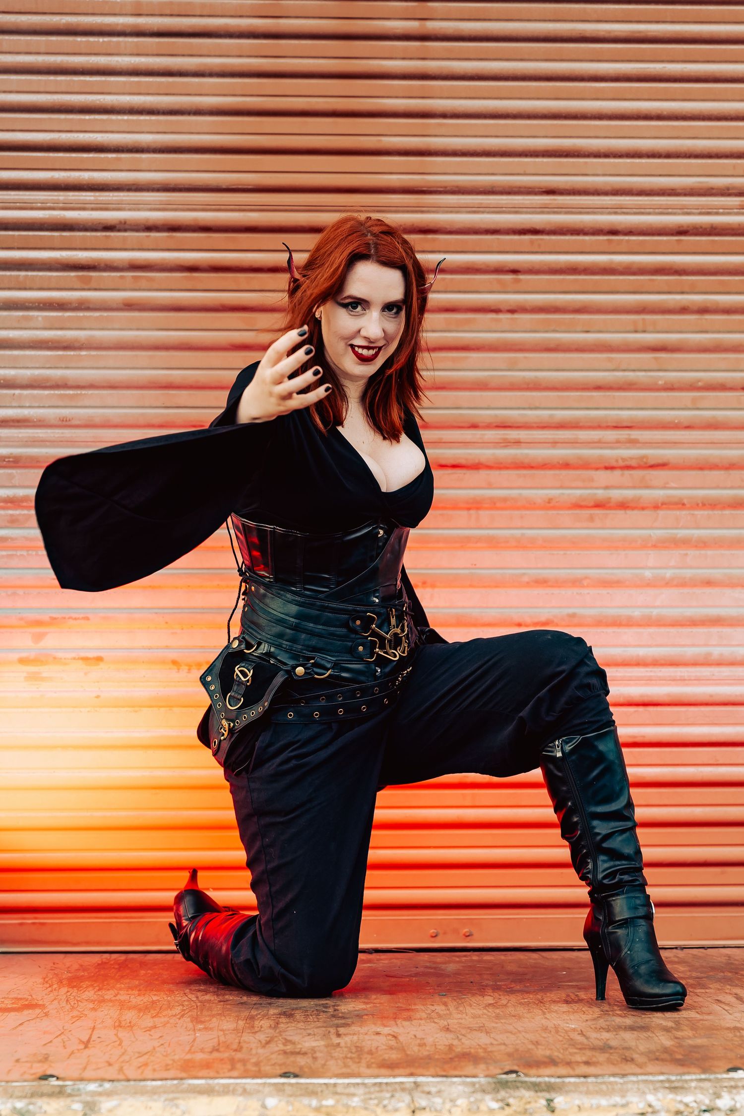 Redhead woman in fantasy cosplay costume with corset and boots kneels dramatically before orange metal backdrop.