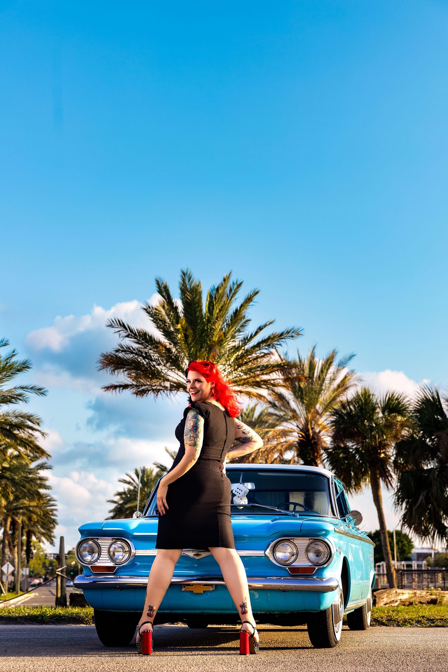 Red-haired woman in black dress and heels poses before a classic blue Chevrolet with palm trees under a sunny sky.