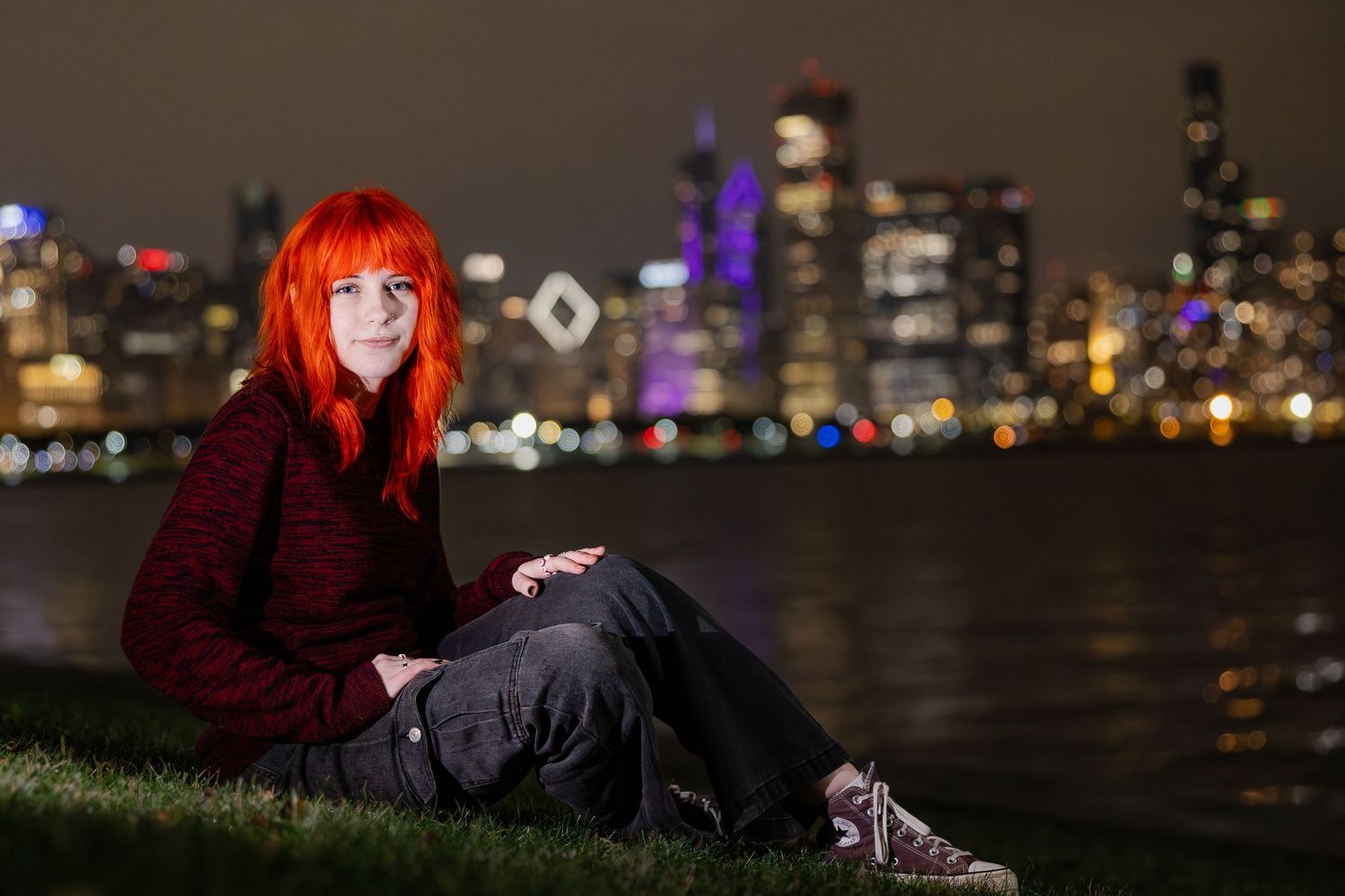 Woman with vibrant red hair sits on grass by waterfront with illuminated city skyline at night.