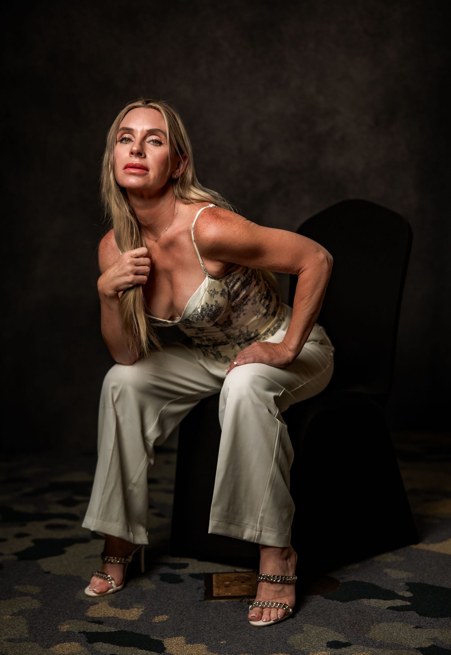 Woman in floral top and white pants seated on black chair, posing confidently against dark studio background.