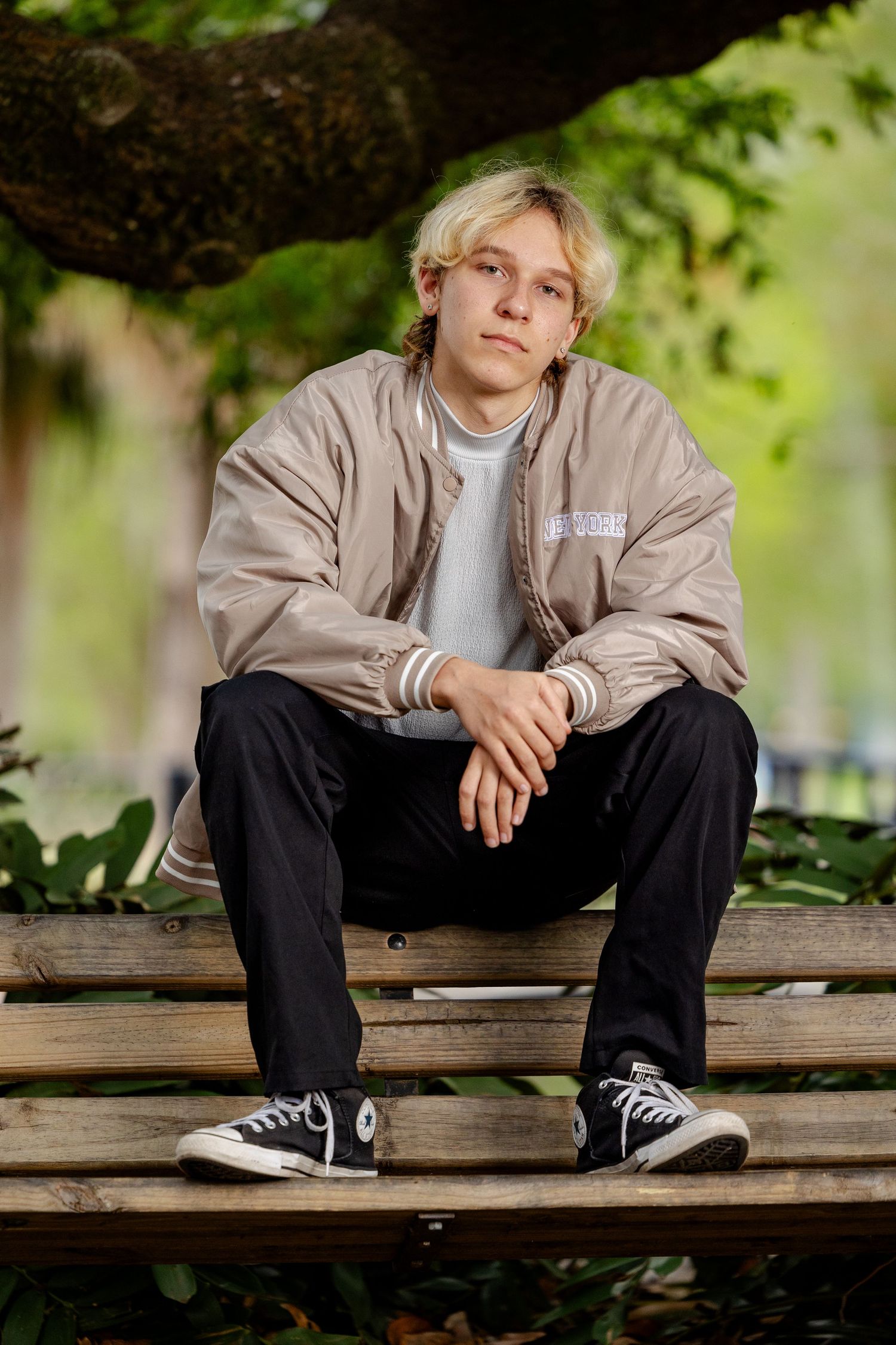 Young man with blonde hair sitting on a wooden bench outdoors, wearing a beige bomber jacket and black Converse sneakers.