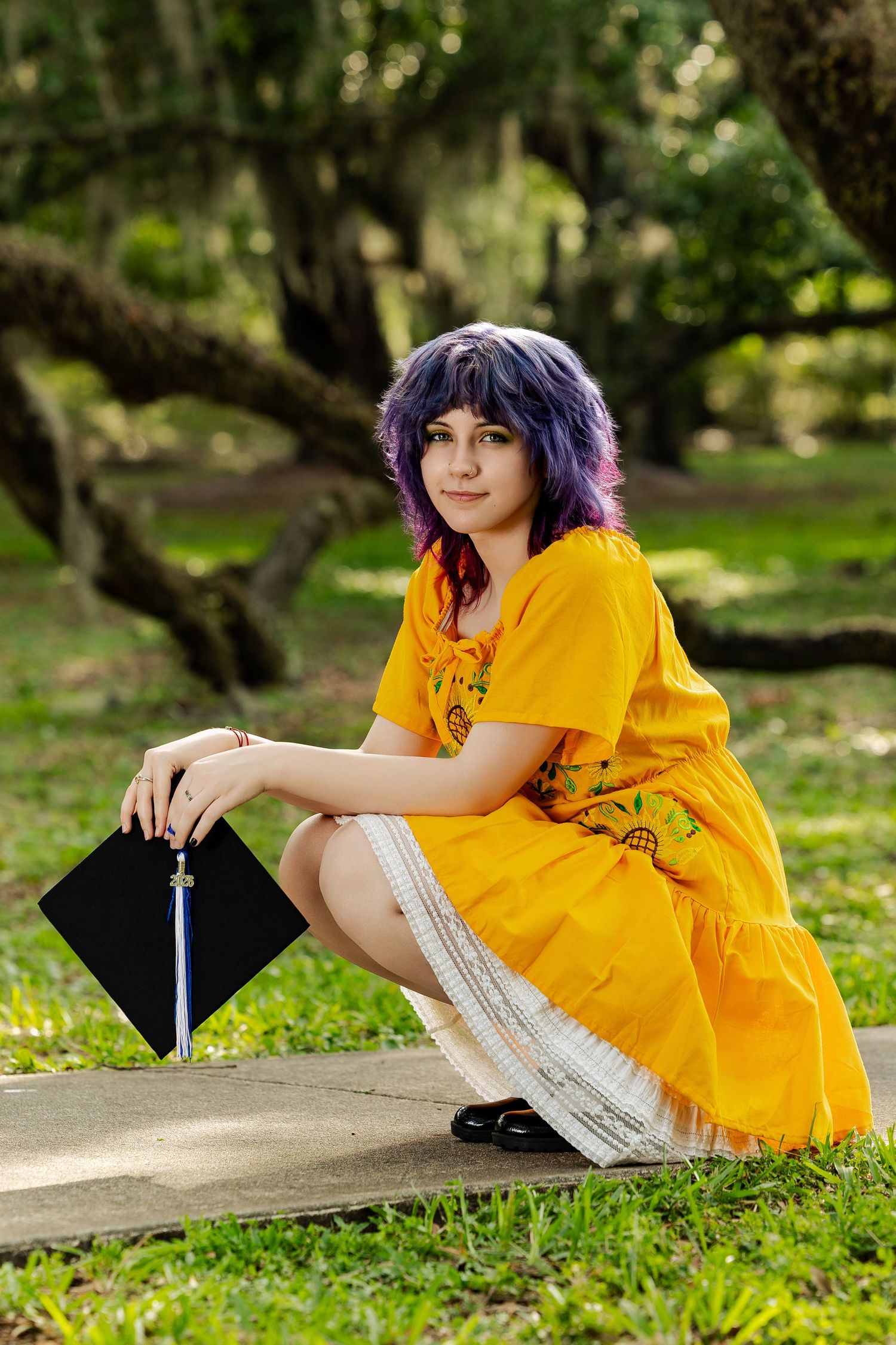 Young woman with purple hair in yellow dress holds graduation cap while sitting outdoors in a lush green park.