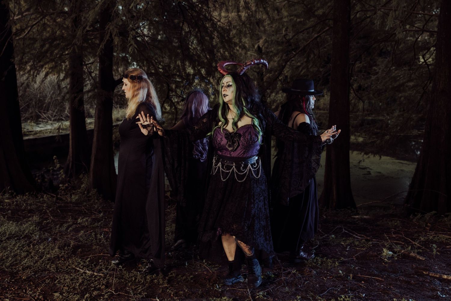 Four people in dark witch costumes pose in a shadowy forest with dramatic gestures for Halloween.