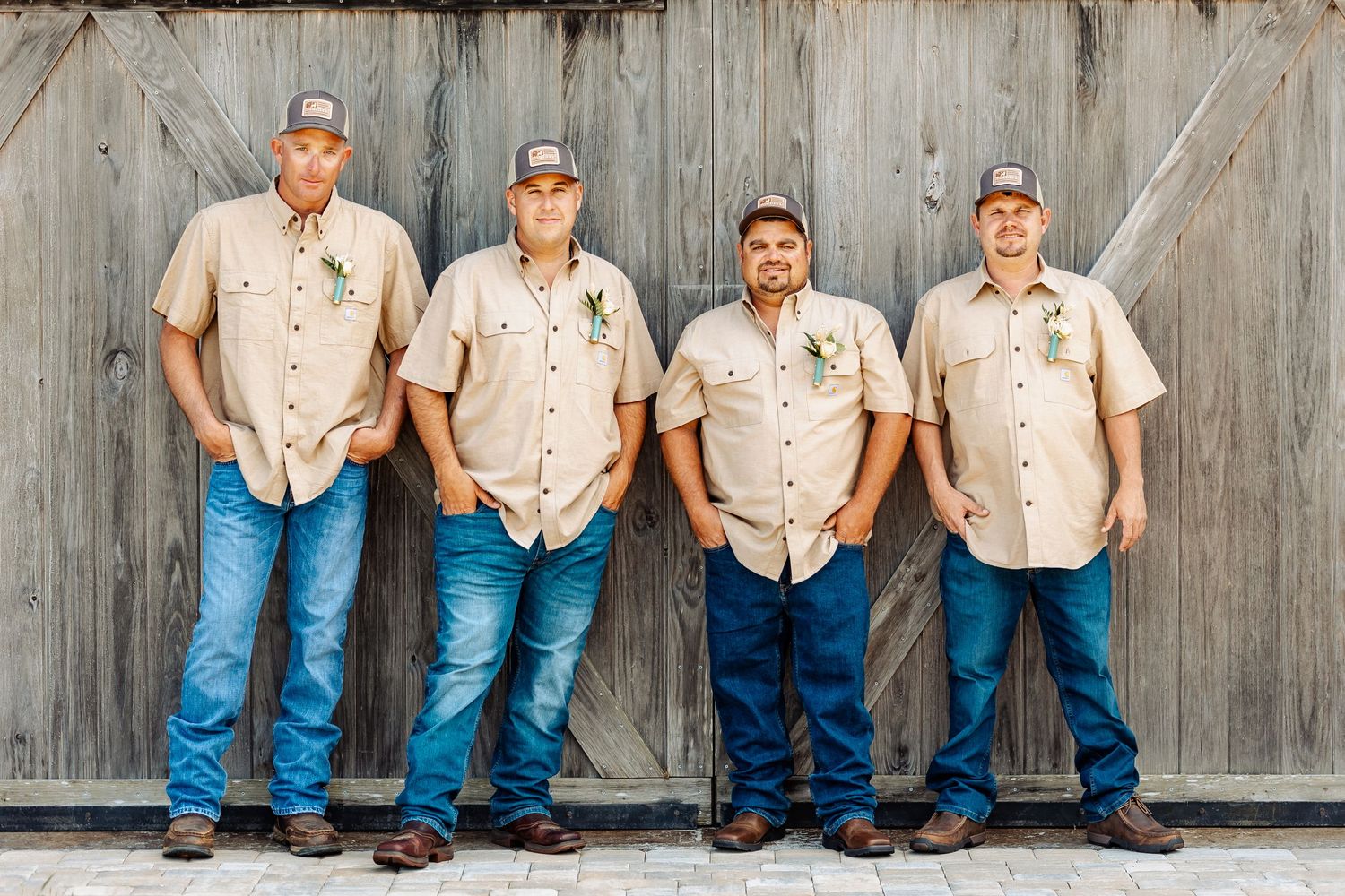 Group wearing matching tan shirts and baseball caps pose against weathered wooden wall.