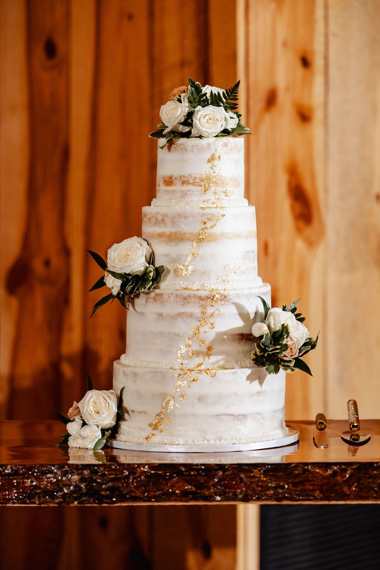 White tiered wedding cake with gold accents and fresh white roses against wooden backdrop.
