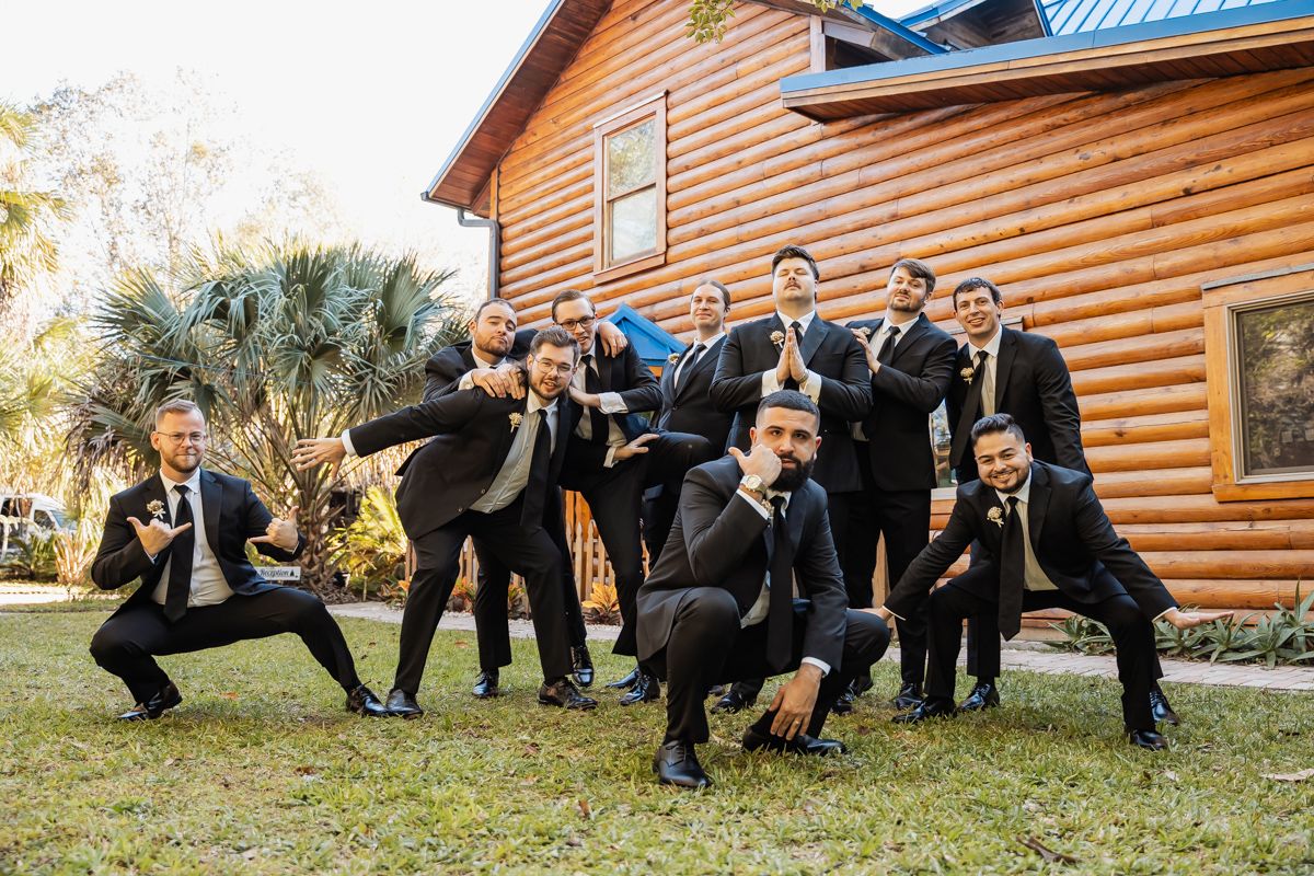 Group of groomsmen in black suits posing playfully outside a rustic log cabin venue.
