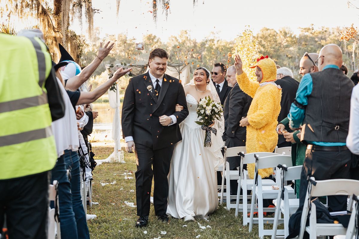 Newlyweds walk down aisle as guests celebrate, one wearing yellow chicken costume, outdoors.