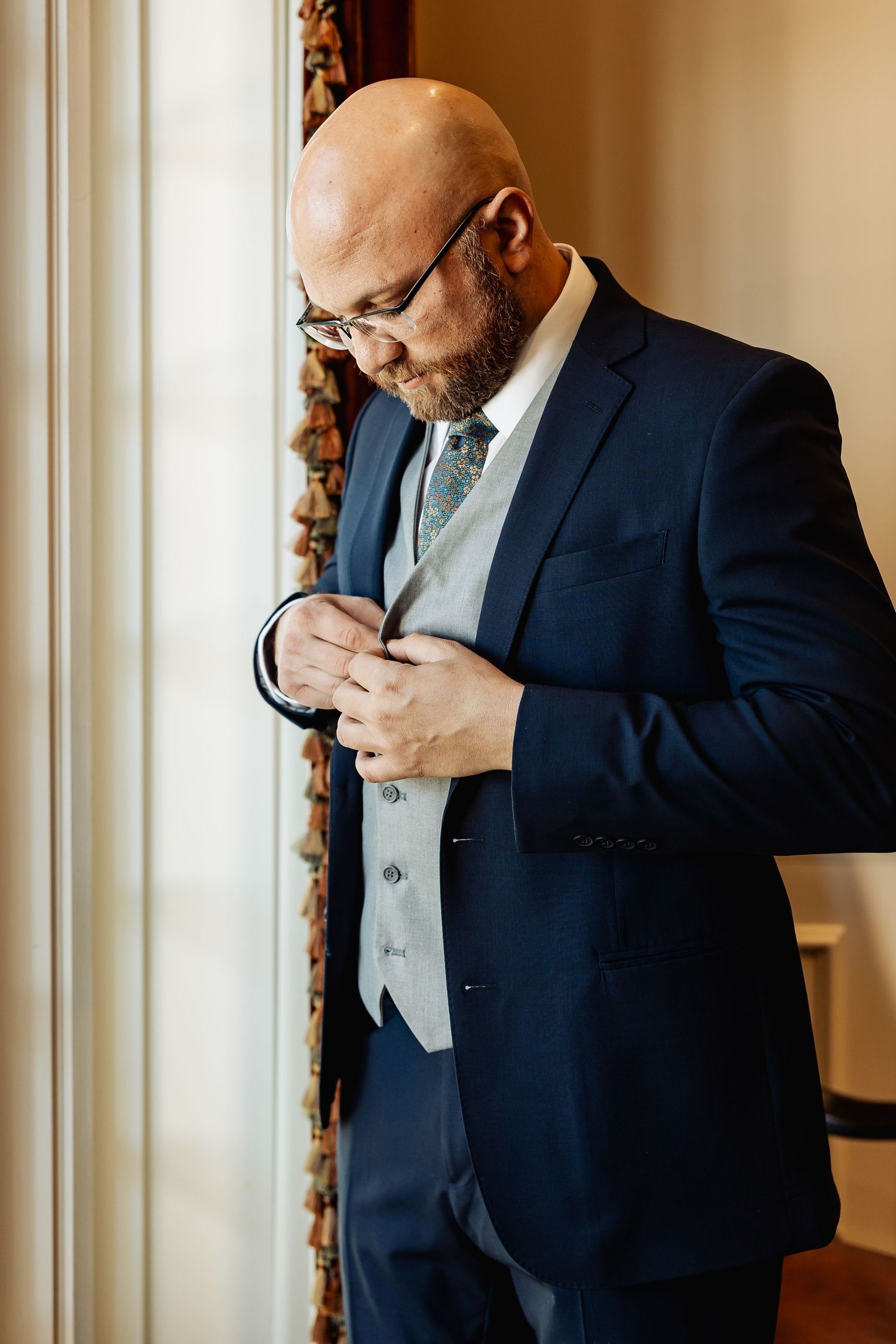 Bald groom in navy suit and gray vest buttoning his jacket near a window.