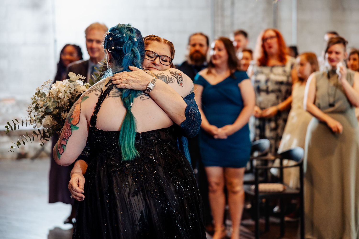 Tattooed bride with teal hair shares emotional hug with bridesmaid in black dress at wedding.