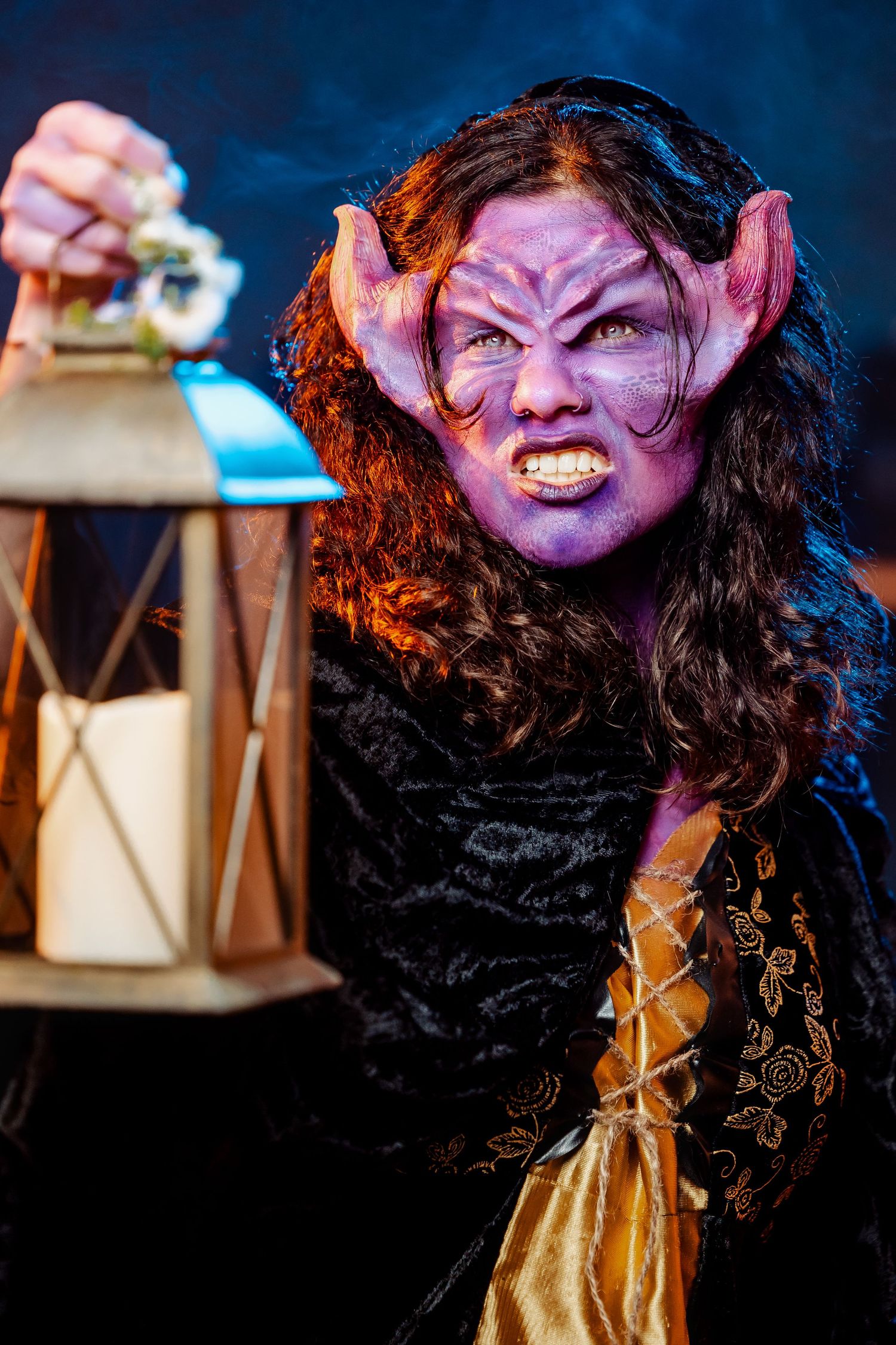 Creature character in black and gold costume holding antique lantern with dramatic lighting.