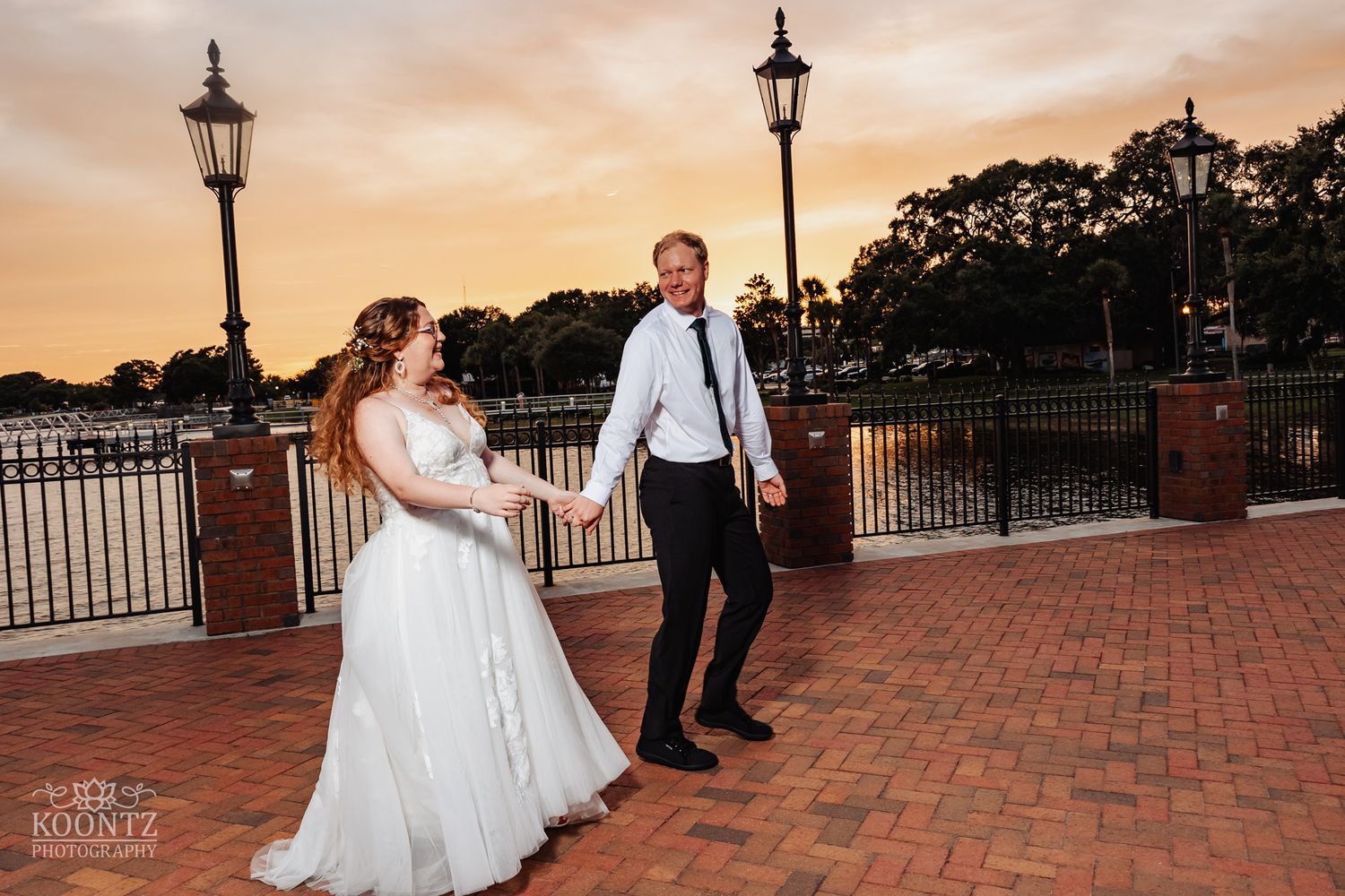 Romantic sunset portrait on a brick walkway with vintage lampposts and waterfront views.