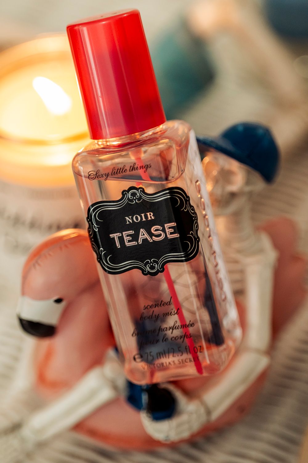 A bottle of 'Noir Tease' perfume with a red cap photographed next to a glowing candle.