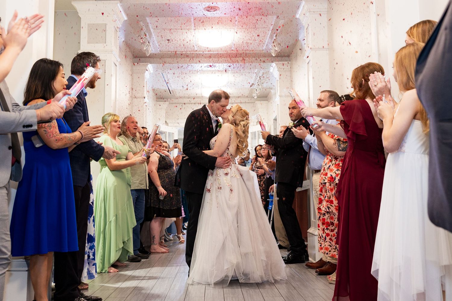 Bride and groom share a kiss during grand exit with confetti and cheering guests lining the aisle.