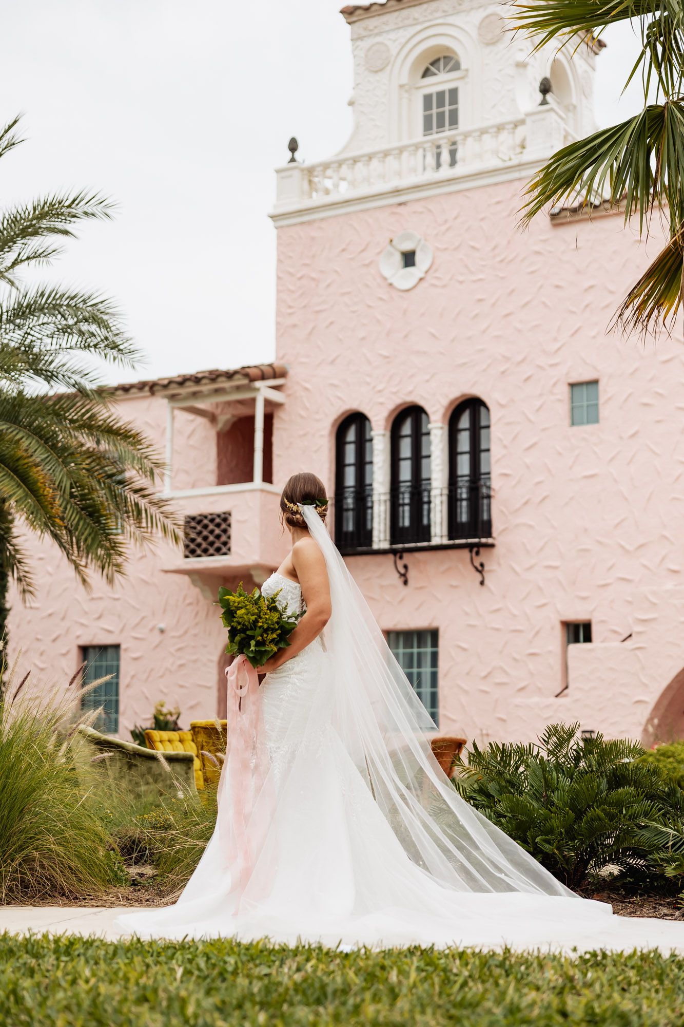 Bride in fitted white gown with cathedral veil stands before a pink Spanish-style wedding venue with palm trees.