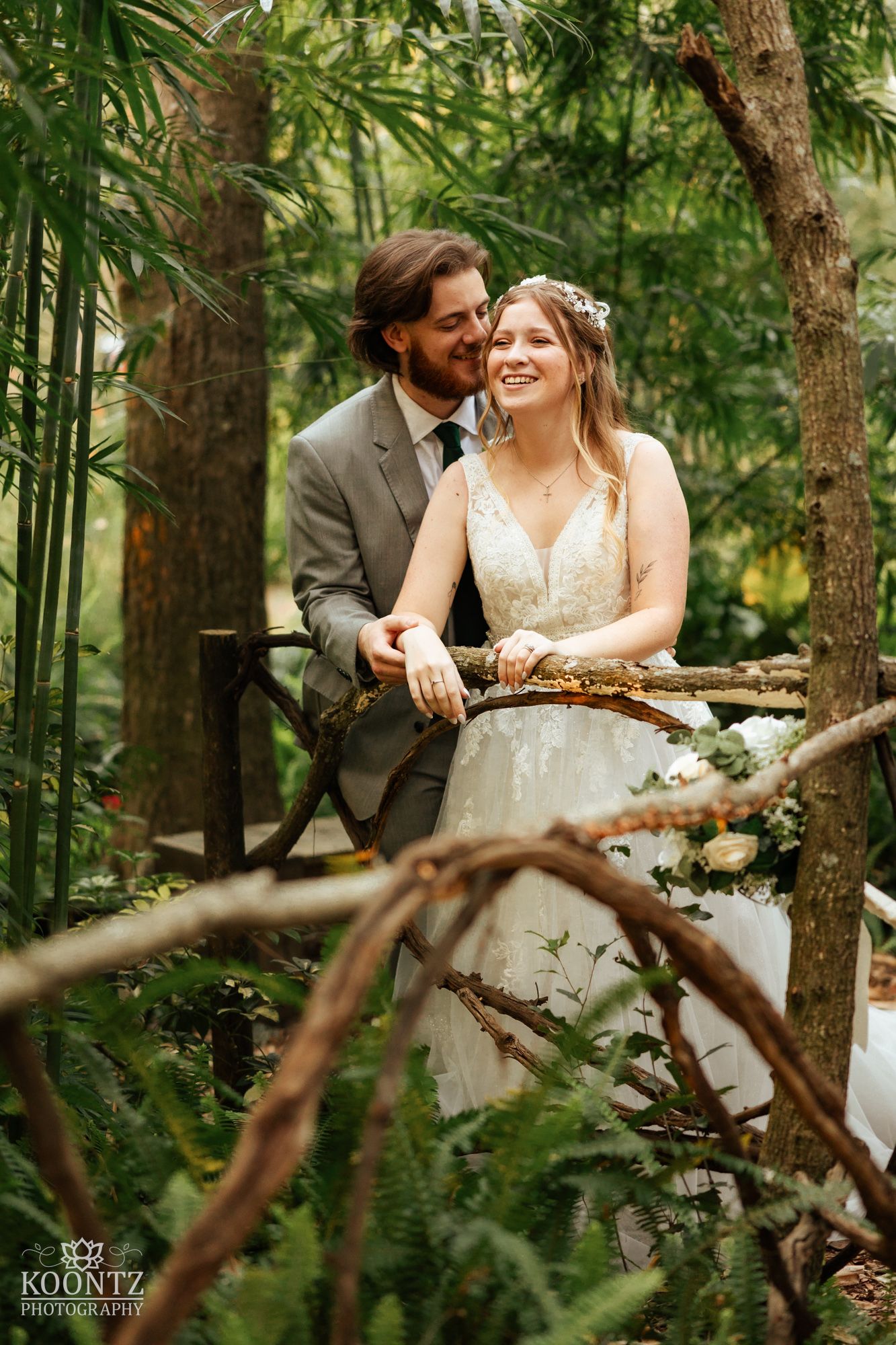 Smiling bride in lace gown and groom in gray suit share tender moment in lush garden setting.