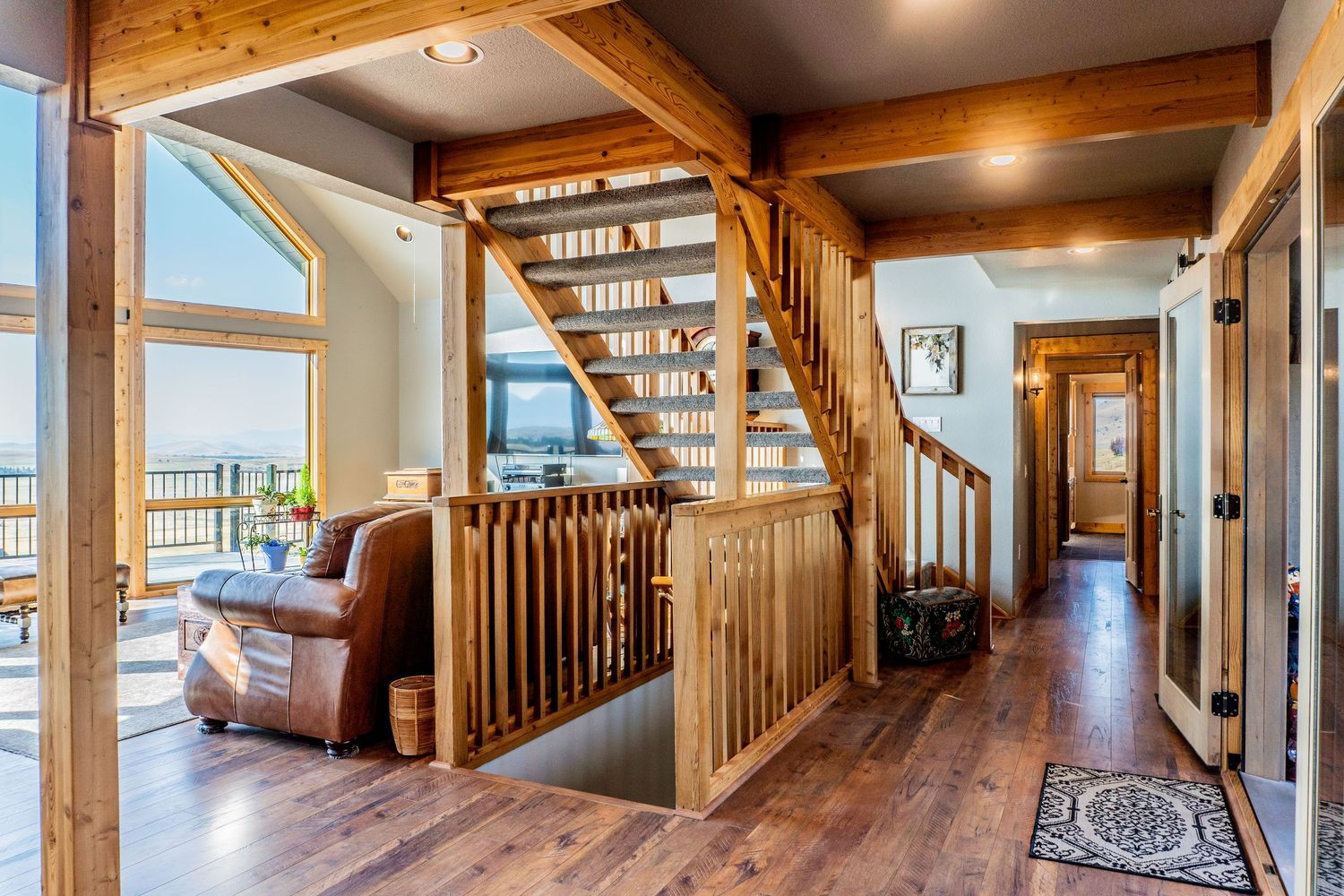 Rustic wooden staircase with log beam accents in an open-concept home featuring hardwood floors and large windows.