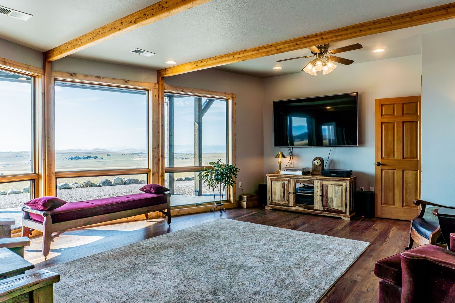 Bright living room with large windows, scenic views, ceiling fan, wall-mounted TV, area rug, and wood-trimmed decor.