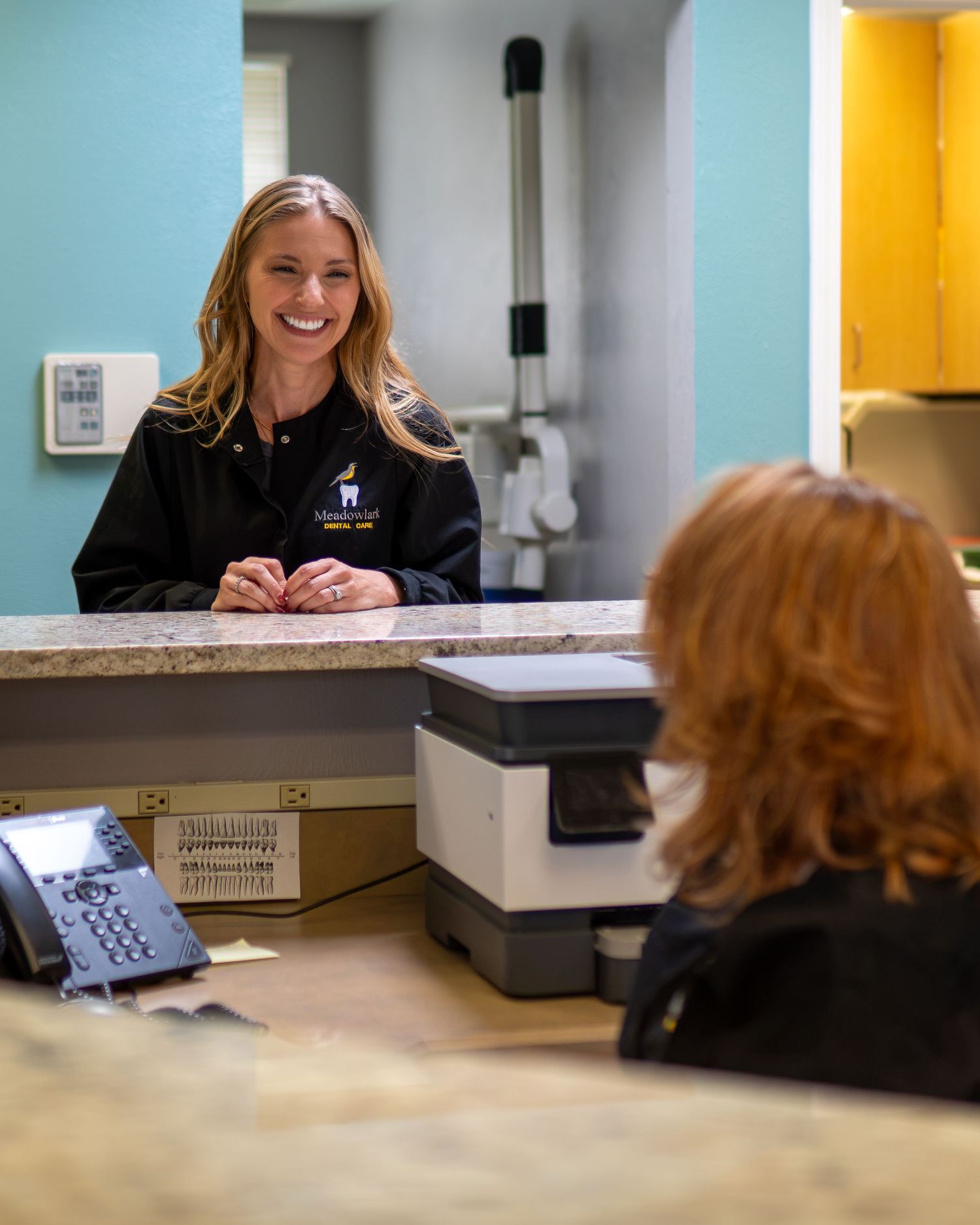 Smiling dental receptionist in black uniform greets a red-haired patient at the front desk of a dental office.
