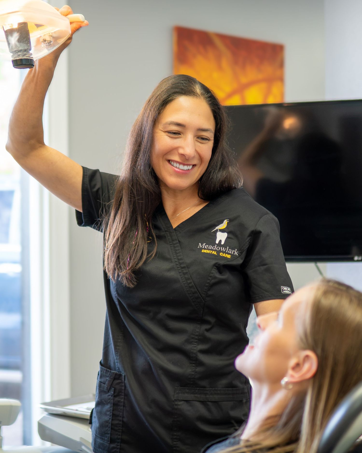 A smiling dental professional in black scrubs holds up a dental model while consulting with a patient in a modern dental office.