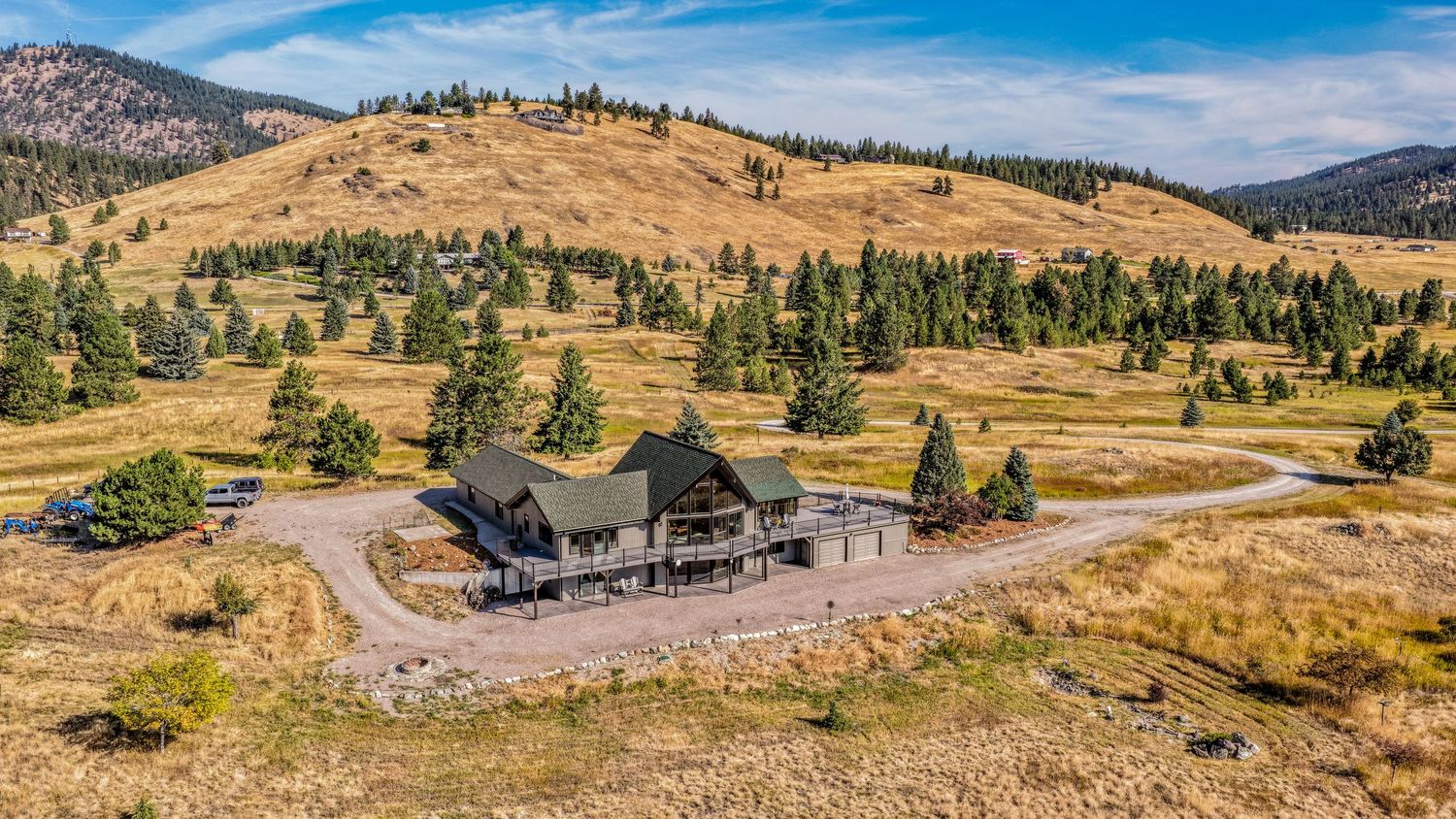Aerial view of a large mountain lodge surrounded by pine trees on golden rolling hills under a clear blue sky.