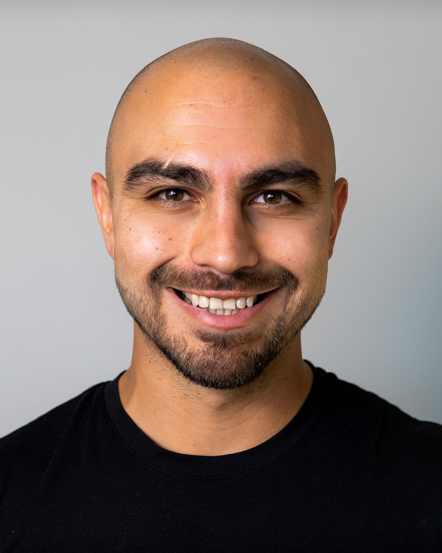 Smiling bald man with beard wearing black shirt, professional headshot on gray background.