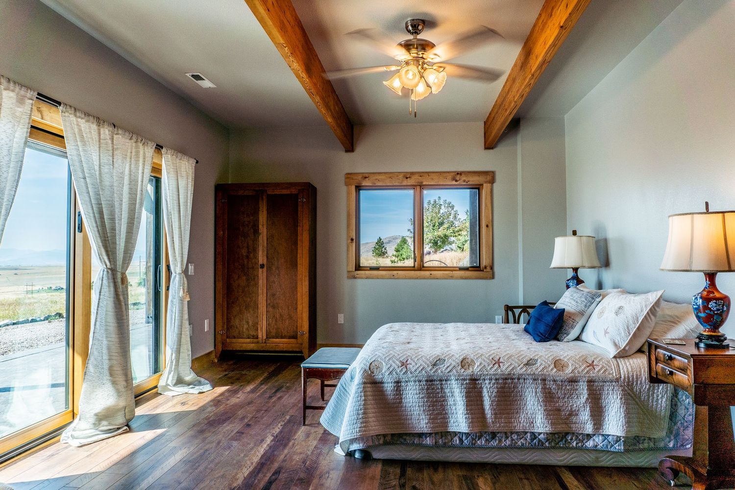 Elegant bedroom with exposed wood beams, hardwood floors, large windows, and rustic wooden armoire.