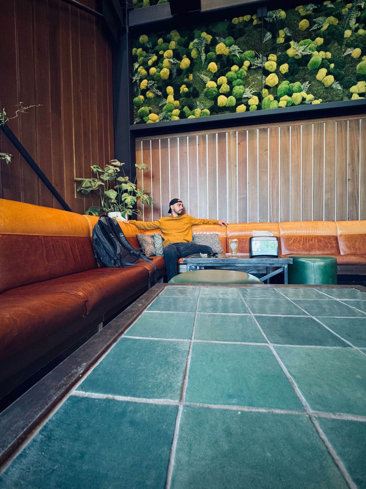 Person in orange sweater relaxing on curved brown leather sofa in retro lounge with green tile floor and plant wall.