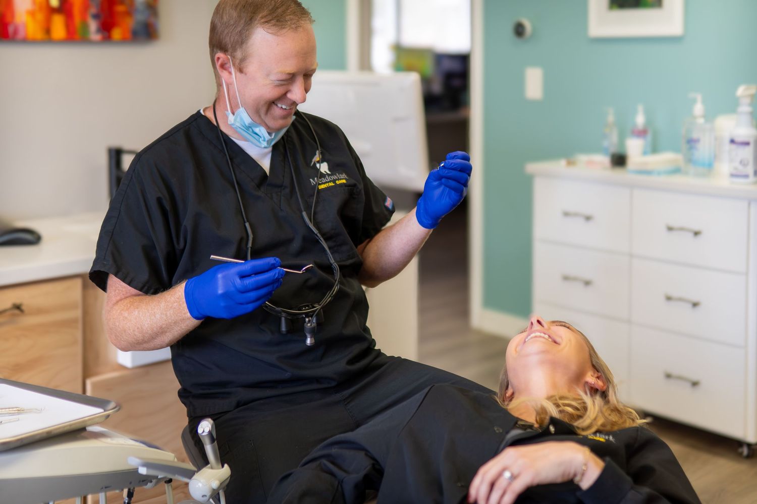 Smiling dentist in black scrubs and blue gloves examining a female patient in a modern dental office.