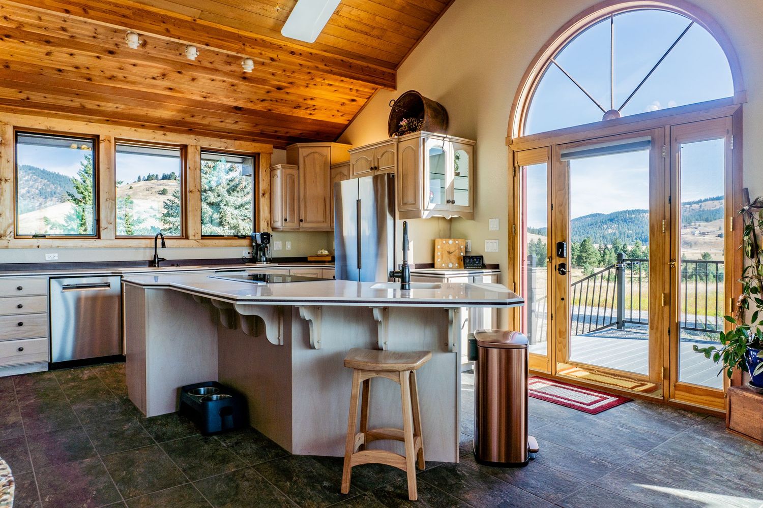 Bright kitchen with wood ceiling, large windows, center island with bar stools, and mountain views beyond French doors.